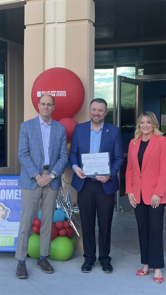 Three people posing in front of a building entrance with a large red Grant a Gift Autism Foundation balloon, one holding a certificate, at the Plaza 8 Henderson ribbon-cutting event